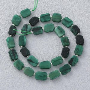 Puede incluir: Un collar de cuentas rectangulares de color verde esmeralda con variaciones en verde oscuro y negro. Las cuentas están ensartadas con un fino cordón verde, probablemente para la fabricación de joyas o manualidades.