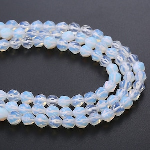 Puede incluir: Una hebra de cuentas de ópalo blanco con un diseño facetado en forma de estrella. Las cuentas están enhebradas en un hilo transparente y están dispuestas en un patrón circular.