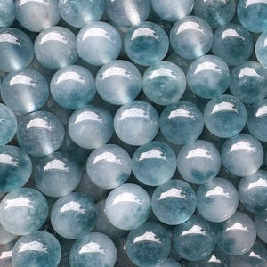 May include: A close-up of a collection of aquamarine beads. The beads are a mix of light and dark blue, with a polished, glossy finish. The beads are strung together, likely for jewellery making or crafts. Each bead is approximately 0.8 cm in diameter.