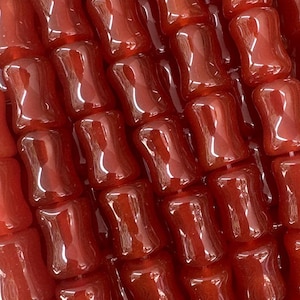 May include: A collection of glossy, reddish-brown beads. Each bead has a unique, elongated shape with a slightly curved design. The beads are strung together, creating a flowing, textured appearance. The beads are held in a hand.