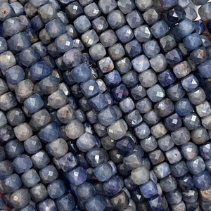 May include: A strand of blue gemstone beads, each bead is faceted and has a square shape. The beads are strung together on a clear thread.