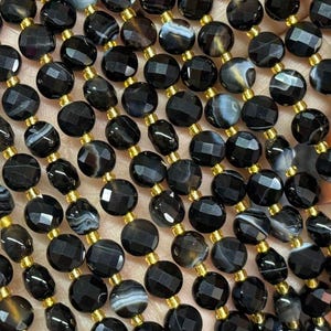Può includere: Una collana di perle di agata nera con accenti dorati. Le perle sono sfaccettate e hanno una forma rotonda. Le perle sono infilate su un filo elastico trasparente.