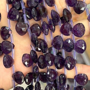 May include: Strands of faceted amethyst beads in various shapes and sizes. The beads are a deep purple color and strung on a clear cord. The beads are held in a hand, showcasing their color and texture.