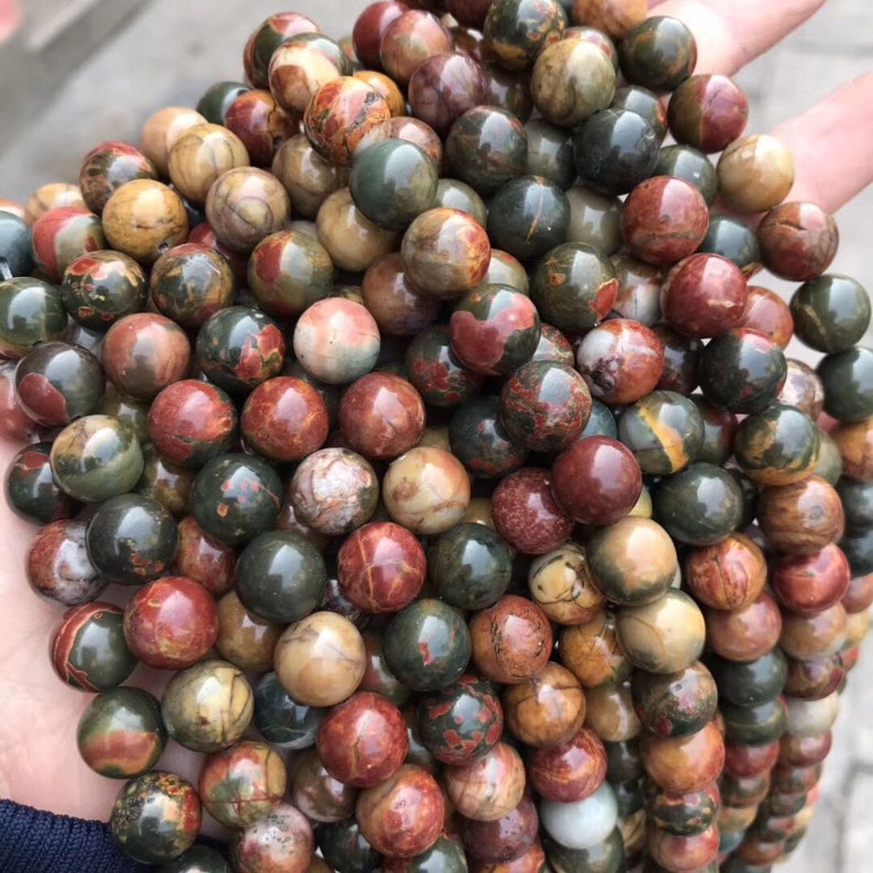 May include: A close-up of a pile of round, multi-colored gemstone beads. The beads are a mix of red, brown, green, and white. The beads are smooth and polished.