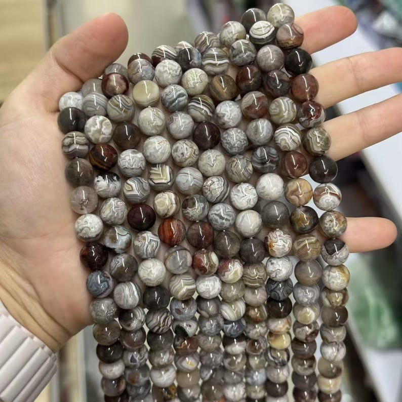 May include: Strands of round gemstone beads in shades of brown, gray, and white. The beads have a marbled appearance, with varying patterns and colors. The beads are held in a hand, showcasing their size and texture. The beads are approximately 8mm in diameter.
