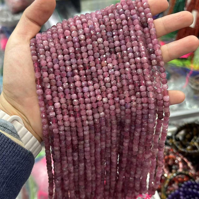 May include: Strands of faceted, square-cut, pink gemstones. The beads are a deep rose color and appear to be strung together. The beads are held in a hand, and the background is blurred.