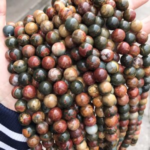 May include: A strand of round, multi-colored gemstone beads. The beads are a mix of brown, red, green, and white. The beads are all about the same size and are strung together on a clear string.