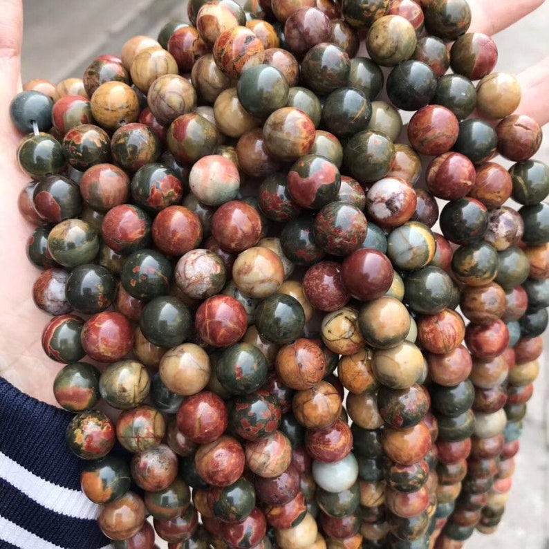 May include: A strand of round, multi-colored gemstone beads. The beads are a mix of brown, red, green, and white. The beads are all about the same size and are strung together on a clear string.