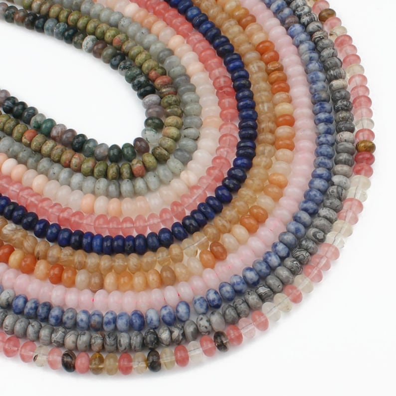 K&ouml;nnte beinhalten: Eine Sammlung von 10 Str&auml;ngen Edelsteinperlen in verschiedenen Farben, darunter rosa, blau, gr&uuml;n und braun. Die Perlen sind rund und glatt und in einem Kreis angeordnet.