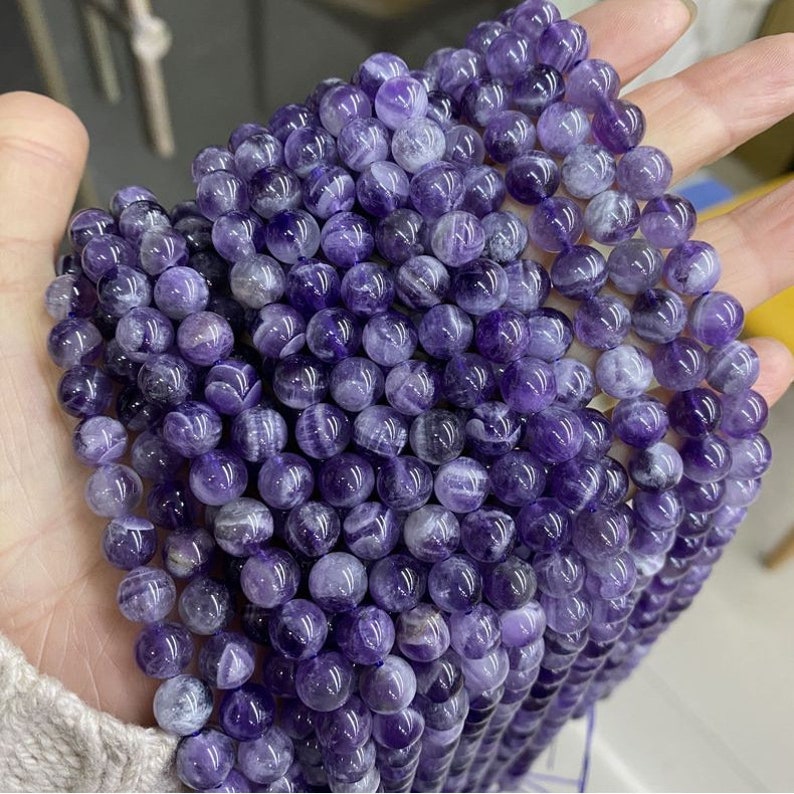 May include: A strand of purple amethyst beads with white streaks. The beads are round and smooth, and they are strung on a clear thread.