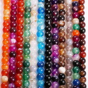 Pode incluir: Conjunto de contas de pedra preciosa de ágata em várias cores, incluindo vermelho, verde, azul, roxo e cinza. As contas são redondas e têm um acabamento liso e polido.