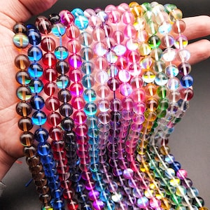 Puede incluir: Una hebra de cuentas de vidrio iridiscente en varios colores, incluidos azul, verde, morado y rosa. Las cuentas son redondas y tienen un acabado liso y pulido.