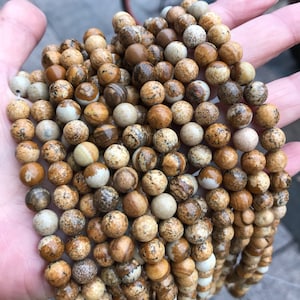 May include: A close-up of a strand of brown and white jasper beads. The beads are round and have a speckled pattern.