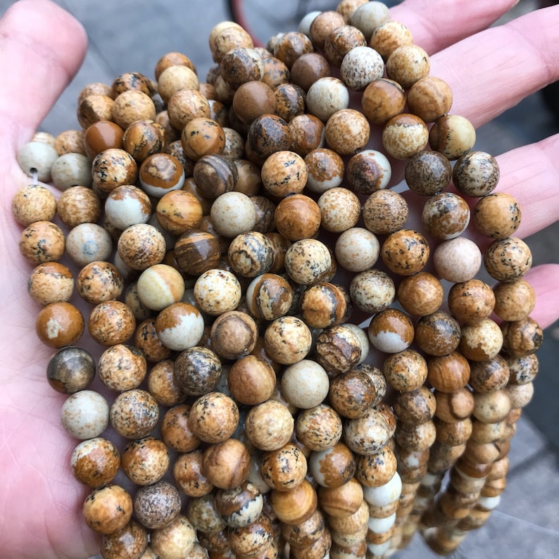 May include: A close-up of a strand of brown and white jasper beads. The beads are round and have a speckled pattern.