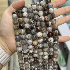 May include: Strands of round gemstone beads in various shades of gray, brown, and cream. The beads have unique patterns and textures, creating a natural and earthy aesthetic. The beads are held in a hand, showcasing their size and shape.