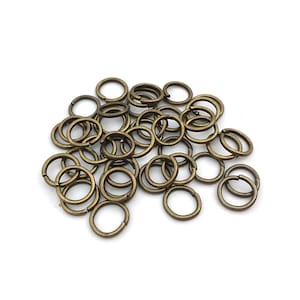 Peut inclure: Un ensemble de petits anneaux fendus de couleur bronze, dispers&eacute;s sur une surface blanche. Les anneaux sont circulaires et fendus, con&ccedil;us pour connecter des composants de bijoux ou d'autres projets d'artisanat. Le m&eacute;tal a un aspect l&eacute;g&egrave;rement vieilli.