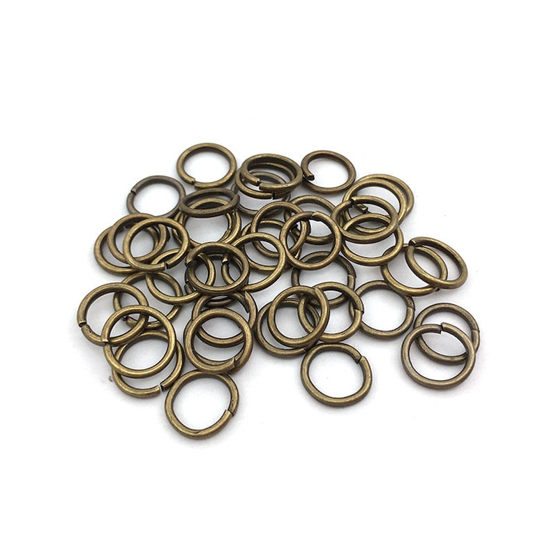 Peut inclure: Un ensemble de petits anneaux fendus de couleur bronze, dispers&eacute;s sur une surface blanche. Les anneaux sont circulaires et fendus, con&ccedil;us pour connecter des composants de bijoux ou d'autres projets d'artisanat. Le m&eacute;tal a un aspect l&eacute;g&egrave;rement vieilli.