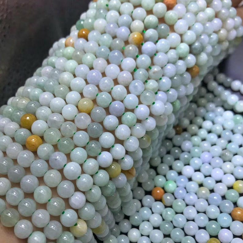 Puede incluir: Primer plano de m&uacute;ltiples hilos de cuentas redondas y pulidas. Las cuentas son principalmente blancas y verde p&aacute;lido, con acentos amarillos y naranjas ocasionales. Las cuentas est&aacute;n ensartadas en un fino cord&oacute;n verde, creando una apariencia delicada y elegante. El tema general es la joyer&iacute;a.