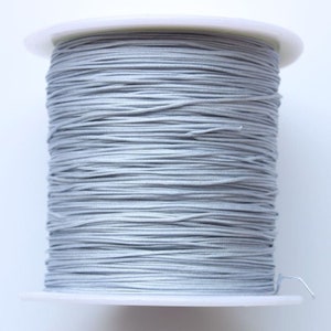 Puede incluir: Una bobina de cord&oacute;n el&aacute;stico plateado, enrollada apretadamente en una bobina de pl&aacute;stico blanca.