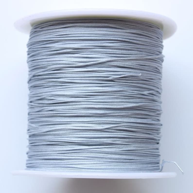 Puede incluir: Una bobina de cord&oacute;n el&aacute;stico plateado, enrollada apretadamente en una bobina de pl&aacute;stico blanca.