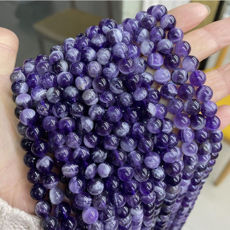 May include: A strand of round, purple amethyst beads with white swirls. The beads are smooth and polished.