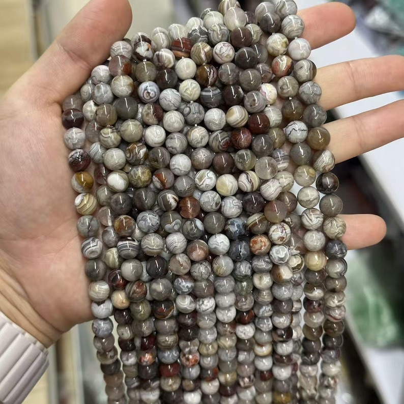 May include: Strands of round gemstone beads in shades of gray, brown, and white. The beads have a marbled appearance and are held in a hand. These beads are suitable for jewelry making.