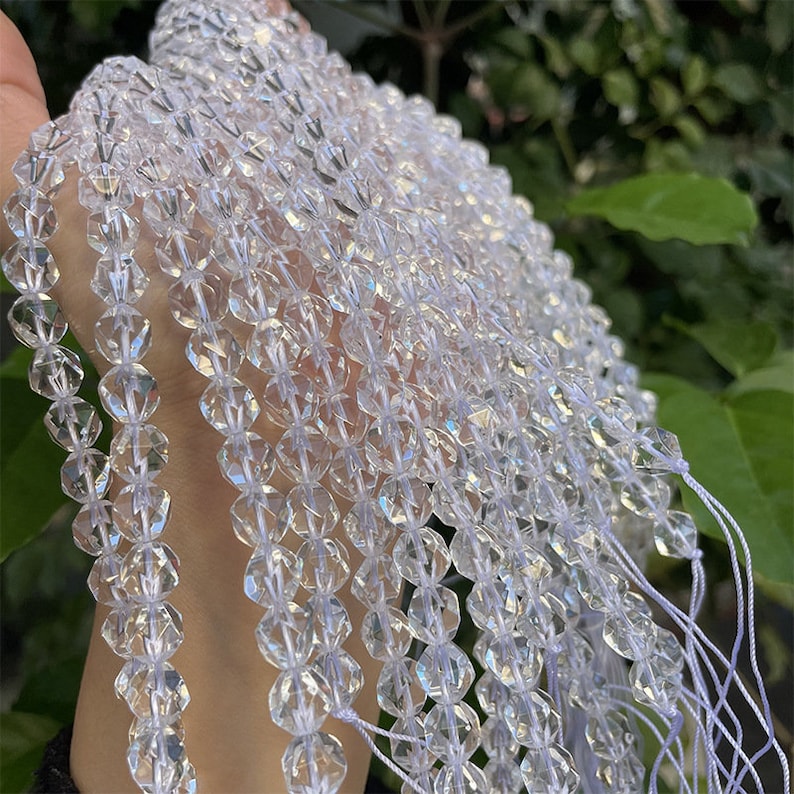 May include: A strand of clear crystal beads, each bead is faceted and has a slightly iridescent quality. The beads are strung on a white thread and are arranged in a cascading pattern.