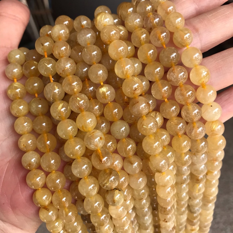 May include: A strand of golden rutilated quartz beads. The beads are round and translucent, with golden inclusions that sparkle in the light.