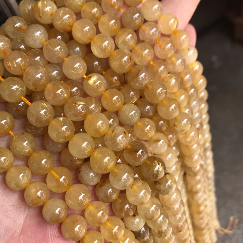 May include: A strand of golden rutilated quartz beads. The beads are round and translucent, with a golden shimmer running through them. The beads are strung on a clear elastic cord.