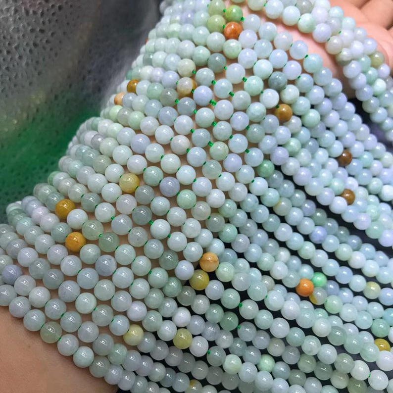 Puede incluir: Una colecci&oacute;n de collares de cuentas de jade. Las cuentas son principalmente de color verde p&aacute;lido con toques de lavanda y amarillo, ensartadas en hilo verde. Las cuentas son redondas y lisas, creando una textura fluida y org&aacute;nica. Los collares se muestran en capas.