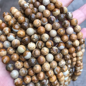 May include: A close-up of a strand of brown and white jasper beads. The beads are round and have a speckled pattern.