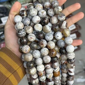 May include: A strand of round gemstone beads in shades of brown, gray, and cream. The beads have unique patterns and textures, and are held in a hand. The beads are approximately 0.4 inches in diameter.