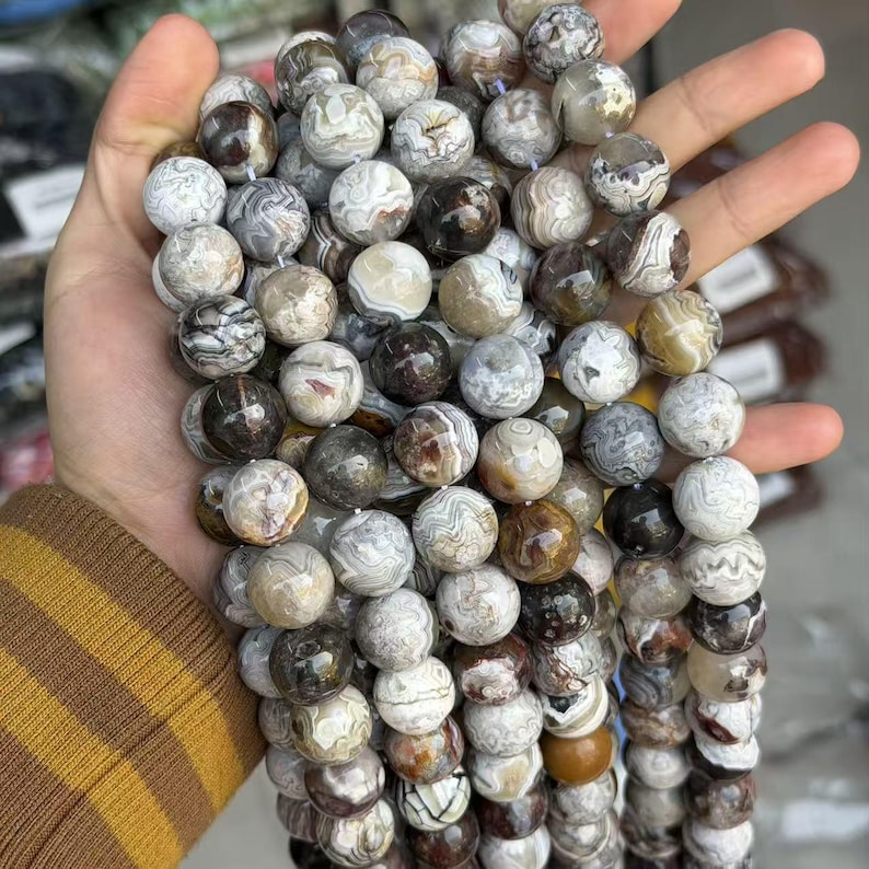 May include: A strand of round gemstone beads in shades of brown, gray, and cream. The beads have unique patterns and textures, and are held in a hand. The beads are approximately 0.4 inches in diameter.