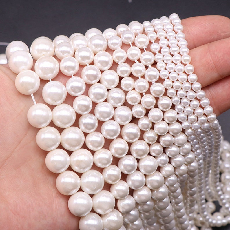 Puede incluir: Una colecci&oacute;n de cuentas de perlas blancas de diferentes tama&ntilde;os, dispuestas en filas sobre una mano. Las cuentas son lisas y brillantes, con un aspecto cl&aacute;sico de perla.