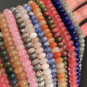 K&ouml;nnte beinhalten: Sortiment an bunten Edelsteinperlen, darunter rosa, blau, gr&uuml;n, orange und wei&szlig;. Die Perlen sind rund und glatt und in Reihen auf einer Hand angeordnet.