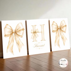 May include: Three rectangular wall art prints featuring watercolour-style illustrations of beige bows. The centre print displays a large letter H adorned with a small bow and the name Harriet written in cursive script below the letter.