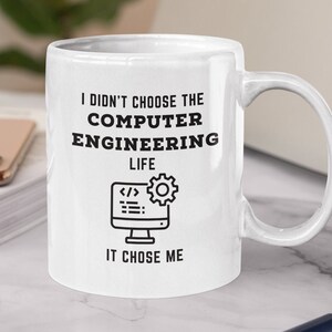 May include: White ceramic mug with black text that reads "I didn't choose the computer engineering life, it chose me". The mug has a black and white illustration of a computer monitor with code and a gear.