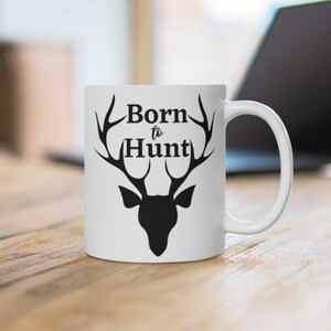 May include: White ceramic mug with a black silhouette of a deer head with antlers. The text "Born to Hunt" is printed on the mug.