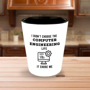 May include: White shot glass with a black rim. It has a black and white graphic of a computer screen with code and a gear. The text reads "I didn't choose the computer engineering life, it chose me."