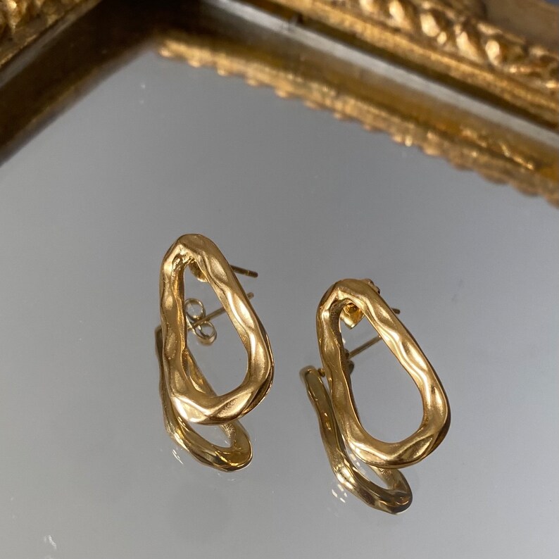 May include: A pair of gold-toned earrings with an irregular oval shape. The earrings have a textured surface and a simple design.