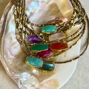 Peut inclure: Plusieurs colliers de pierres précieuses avec des chaînes dorées. Chaque collier présente une pierre précieuse rectangulaire de couleur différente, notamment verte, violette, rouge et bleue. Les colliers sont exposés sur une surface nacrée.
