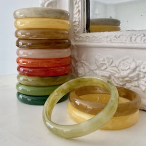 Op de afbeelding: Een stapel kleurrijke plastic armbanden in tinten groen, geel, oranje en bruin. De armbanden zijn gestapeld, met de grootste armband onderaan en de kleinste bovenaan.