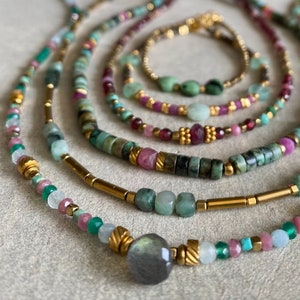 May include: A layered necklace with multiple strands of beads in shades of green, pink, and gold. The necklace features a large, grey gemstone pendant.