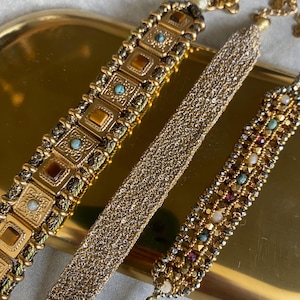 Peut inclure: Trois bracelets dorés aux motifs complexes sont présentés sur un plateau doré. L'un présente des pierres ambrées carrées et des accents turquoise. Un autre est un maillage de petites perles dorées. Le troisième a un design perlé avec diverses pierres colorées.