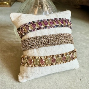May include: Three bracelets displayed on a white pillow. The top bracelet features purple gemstones, the middle one is a gold chain, and the bottom one has green and pink gemstones. The bracelets are arranged on a white pillow.