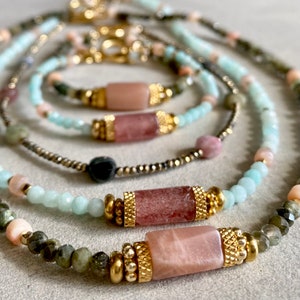 Può includere: Un set di collane sovrapposte con perline di pietre preziose di vari colori, tra cui verde, rosa e blu. Le collane sono decorate con perline di metallo dorato e pietre rettangolari rosa.