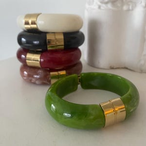 May include: A stack of five colorful bangles. The bangles are in cream, black, burgundy, brown, and green. Each bangle has a gold-colored clasp. The green bangle is in the foreground, and the other bangles are stacked behind it.