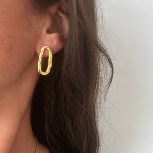 May include: Gold oval-shaped earrings with a textured surface. The earrings are worn in a person's ear.