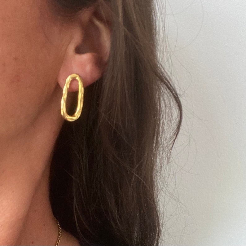 May include: Gold oval-shaped earrings with a textured surface. The earrings are worn in a person's ear.