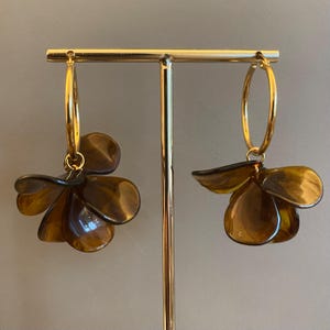 May include: Gold hoop earrings featuring brown, flower-shaped pendants. The earrings are presented on a gold stand. The flower petals have a translucent, glossy finish. The background is a neutral colour.